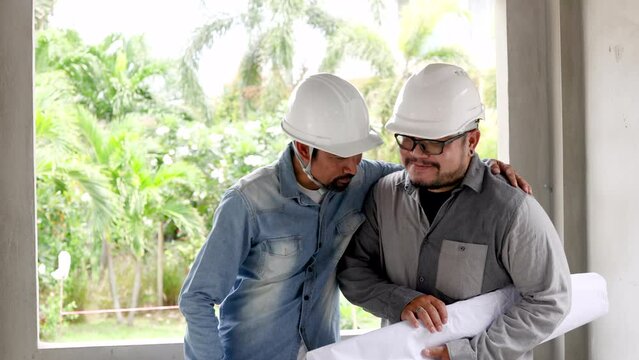 Senior middle-aged Indian engineer admire Asian foreman with mustache wearing safety helmet hardhat medium shot, architect advisor pat shoulder young employee male hold blueprints at construction site