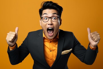 Surprised young Asian businessman showing thumbs up gesture over yellow background