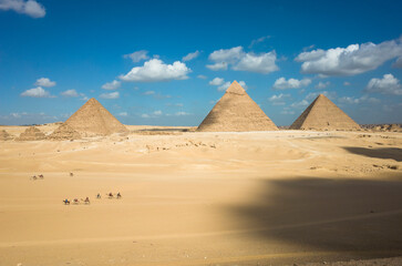 Fototapeta na wymiar Giza pyramid complex in Egypt, The three main pyramids and small subsidiary pyramids, Camel caravan with tourists at sightseeing, White clouds on blue sky, Shadow from clouds on desert yellow sand