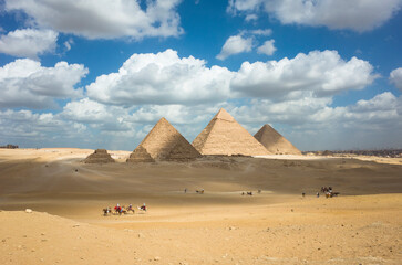 Giza pyramid complex in Egypt, The three main pyramids together with subsidiary pyramids, Tourists sightseeing on camels and horses, White clouds on blue sky, Shadow from clouds on desert yellow sand