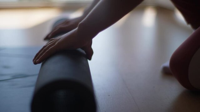 Close-up cropped shot of unrecognizable preteen girl rolling up yoga mat after training at home by window. Closeup hands of child girl folding fitness mat on floor at yoga studio, slow motion.