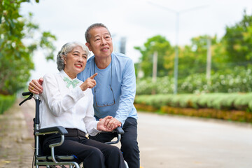 Fototapeta na wymiar Mature asian female people sitting on wheelchair is closely monitored by her husband. elderly couple at park outdoor, relax, elderly caregiving, husband devotion to wife in park setting