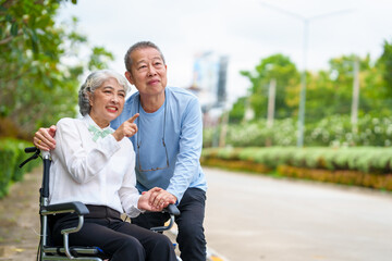 Mature asian female people sitting on wheelchair is closely monitored by her husband. elderly couple at park outdoor, relax, elderly caregiving, husband devotion to wife in park setting