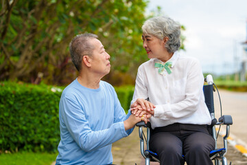 Mature asian female people sitting on wheelchair is closely monitored by her husband. elderly couple at park outdoor, relax, elderly caregiving, husband devotion to wife in park setting