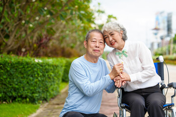 Mature asian female people sitting on wheelchair is closely monitored by her husband. elderly couple at park outdoor, relax, elderly caregiving, husband devotion to wife in park setting