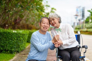 Mature asian female people sitting on wheelchair is closely monitored by her husband. elderly couple at park outdoor, relax, elderly caregiving, husband devotion to wife in park setting