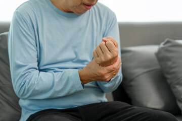 Elderly Asian gentleman with wrist discomfort on the couch, expressing pain due to Carpal Tunnel...