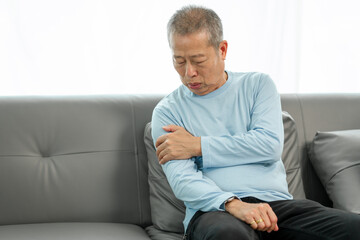 Fototapeta premium Senior Asian man suffering from intense shoulder pain on the sofa, in need of comfort. Painful moment