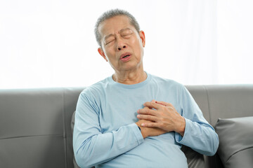 Elderly asian man on sofa, clutching chest in severe pain, showing signs of possible heart attack at home. serious health concern, home emergency, health crisis, chest pain alarm