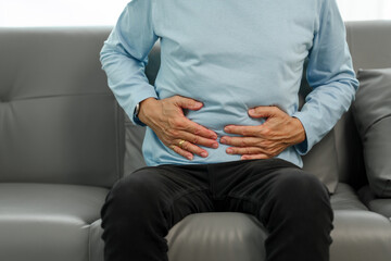 Stomach agony, Elderly Asian man sitting on the sofa, enduring severe pain and discomfort in his abdominal area, clutching his stomach in pain