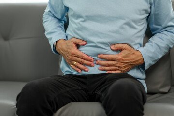Stomach agony, Elderly Asian man sitting on the sofa, enduring severe pain and discomfort in his abdominal area, clutching his stomach in pain