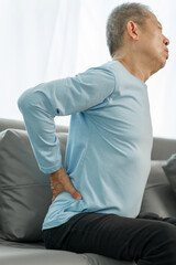 Elderly Asian man on sofa, grimacing from spinal pain, seeking relief from osteoporosis-related discomfort at home.
