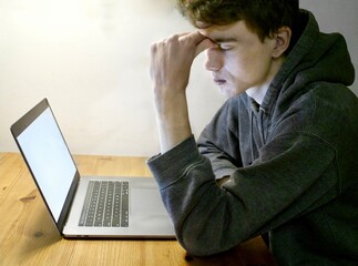 A young man sits in front of a laptop, with his eyes closed and pinching his furrowed brow in frustration.