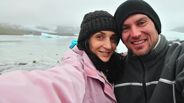Happy multiethnic couple taking selfie by glacier lagoon in Iceland. Two tourists having fun on romantic winter vacation in Iceland - Holidays and traveling lifestyle concept