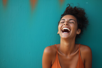 Black beautiful model teenage woman with curly hair dressed in orange tak top laughing and smiling in front of a teal wall, funny, happiness, joy, enjoy life - obrazy, fototapety, plakaty