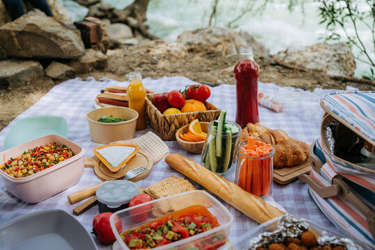 Picnic in the forest camping site with vegetables, juice, cheese, and croissants near mountain river. Fresh organic veggies surrounded with bread baguettes, salads.