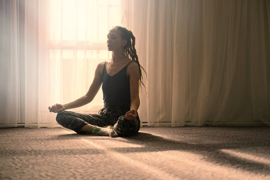 Woman sitting in meditative pose in front of large window at home
