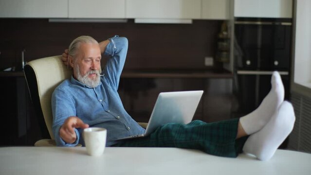 Confident mature aged businessman typing on laptop, putting feet on table in home office room, drinking coffee on workplace. Happy older-aged bearded male working on laptop enjoying beverage.