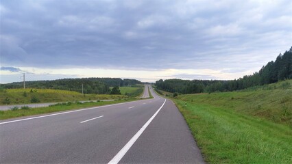 An asphalt road with markings and a dividing strip runs among grassy meadows. There are trees and power poles along the road. The sky is covered with clouds