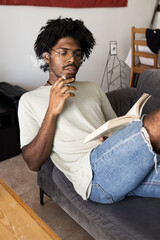 Afro boy with curly hair smoking on the sofa at home while reading a book. Vintage vertical photo