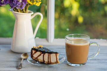 Cheesecake with chocolate sauce and glass cup of cappuccino coffee on a wooden table near window, closeup. Coffee time with slice of cake at morning