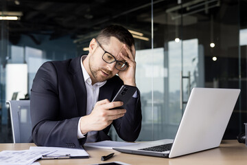 Upset young man businessman, office worker sitting at desk and holding hand on head, got bad news, reading message