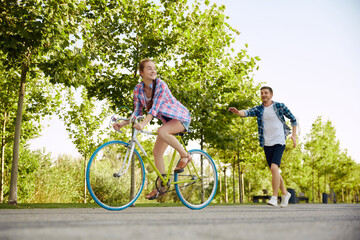 Cheerful active young couple with bicycle in public park together having fun. Man catches up with girlfriend on bike. Happy couple riding cycle