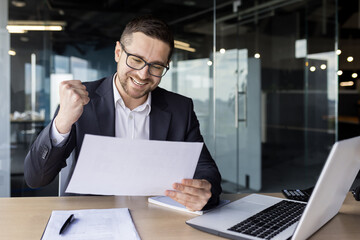 Happy young businessman working in the office, holding documents, agreement, financial profit report in his hands and enjoying success, showing a victory gesture with his hand