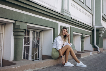 Beautiful blonde model, fashionable in a dress and jacket, poses against the walls of a building