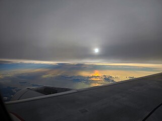 View from the porthole of a flying aircraft from a high altitude on the wing of the aircraft, clouds and the ocean with the setting sun reflected in it
