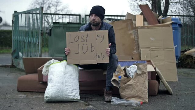 Frozen untidy young man sits by a pile of rubbish and holds a handwritten LOST MY JOB HELP poster.