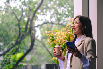 Confidence Asian businesswoman holding smartphone and paper cup, enjoying the fresh air and gentle breeze in the nature