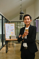 Portrait of millennial businessman holding disposable cup standing in office and looking at camera