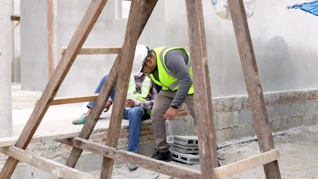 Hopeless middle-aged Indian worker stressed sitting at construction site, young foreman colleagues give bottle of water to laborer man and talking about financial crisis or termination of employment