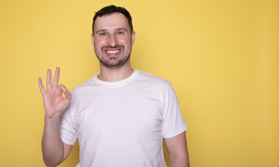 Portrait of handsome excited man in basic clothing smiling and showing ok sign at camera isolated over yellow background.
