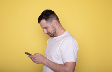 Smiling young bearded man guy in white casual t-shirt posing isolated on yellow background. People lifestyle concept. Using mobile phone, typing sms message.