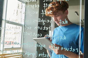 Student using digital tablet while writing computer codes on glass wall at lesson