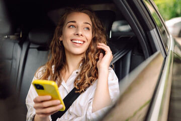 Beautiful young woman uses a smartphone while sitting in the back seat of a car. Concept of technology, traveling by car, business.