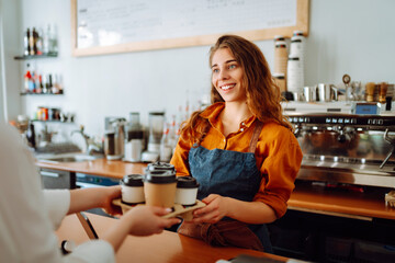Takeaway food concept. A beautiful female barista gives to-go coffee to a client. Owner of a small business, a coffee shop,  behind the bar. Business concept.