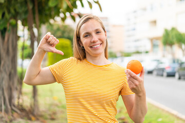 Young blonde woman holding an orange at outdoors proud and self-satisfied