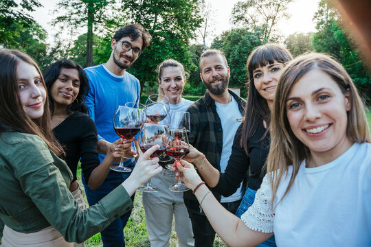 Mixed group of friends take a selfie with smartphone toasting together with glasses of red wine during a party in an outdoor garden at sunset - Happy people having fun in a relaxed moment - Copy space