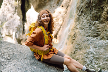 Smiling female traveler with a yellow backpack, dressed in hiking clothes, enjoys a waterfall near a mountain river. Travel, trekking. Nature concept.