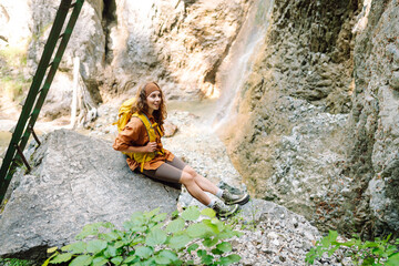 Smiling female traveler with a yellow backpack, dressed in hiking clothes, enjoys a waterfall near a mountain river. Travel, trekking. Nature concept.