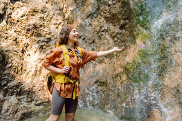 Smiling female traveler with a yellow backpack, dressed in hiking clothes, enjoys a waterfall near a mountain river. Travel, trekking. Nature concept.