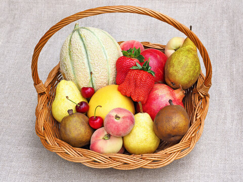 Different fresh colorful fruits in wicker basket on natural fabric background. High angle view of assorted organic berries melon peach pear orange pomegranate  .