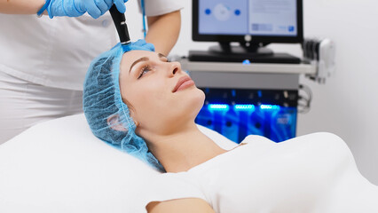 Fototapeta na wymiar Cosmetology clinic. Professional female cosmetologist doing hydrafacial procedure while being a work. Attractive nice woman lying on the medical bed while having beauty procedures