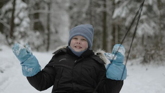 Portrait of happy boy waving his hands in mittens and smiling at camera during sled ride. Winter family walk through snowy forest of positive child with parents and cross narrow road in countryside.