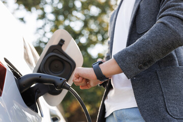 Male hand inserts power connector into EV car and charges batteries, uses smartwatch for activates start charging. Businessman plugging in charging cable to to electric vehicle