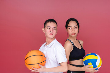 Two young and active players posing back to back while holding a volleyball and a basketball. Isolated on a red background.