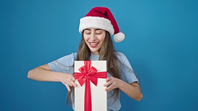 Young beautiful hispanic woman wearing christmas hat holding gift over isolated blue background